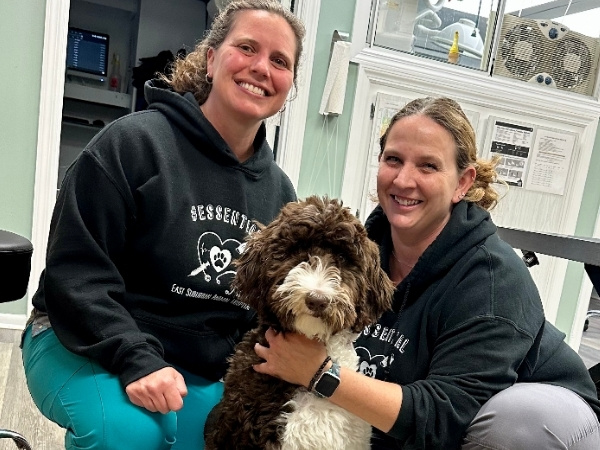 Two people in black hoodies and scrubs kneel on the floor with a brown and white dog between them.