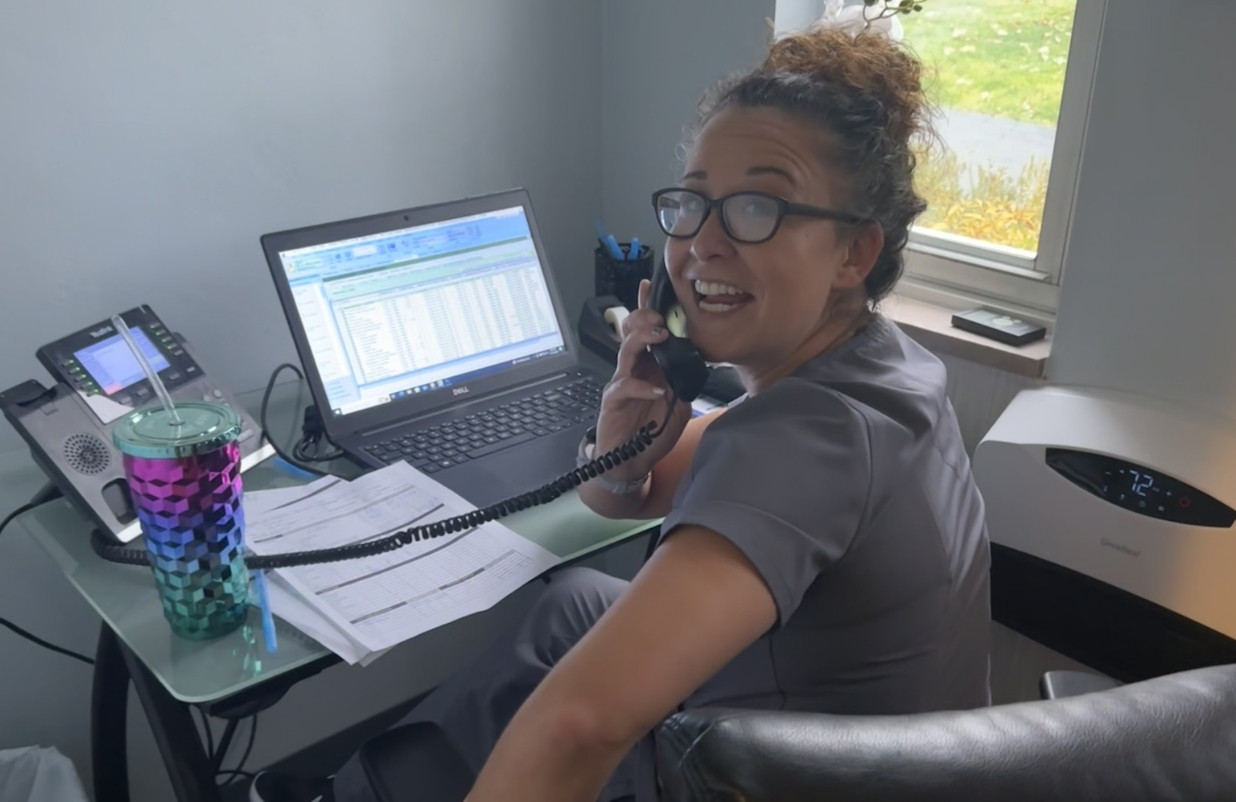 A person sits at a desk talking on a corded phone and looking at a laptop with a spreadsheet open. A colorful tumbler sits on the desk next to a phone and papers.