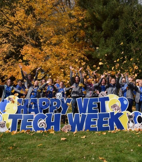 A group of people in front of a "Happy Vet Tech Week" sign outdoors during the fall, with leaves falling around them.